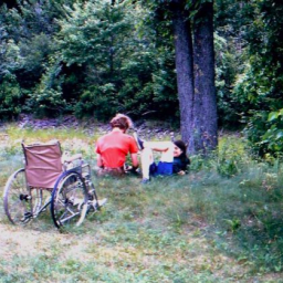 In this faded picture from the 1970s, we see two people sitting by a tree in a grassy field. Behind them is a wheelchair. 