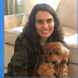 Image of Alexandra Nicklas. Alexandra has dark brown medium length hair. She is wearing a green camouflaged zip-up hooded sweatshirt and tan pants. She is sitting in front of a cream colored sofa. It is daylight, which can be noticed through a nearby window. Ali appears to be at a home setting. Ali is smiling and holding her golden doodle dog, Sadie. 