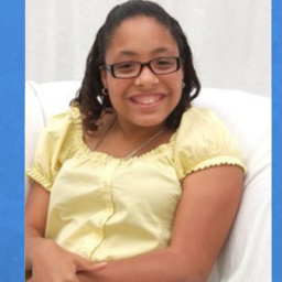 Image of Tylia Flores. Tylia is seated in a plush white chair with a white background. She is wearing a pale-yellow short sleeve blouse. Tylia has dark skin and medium length black hair. She is wearing glasses and smiling at the camera..